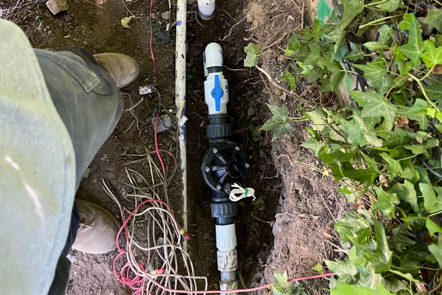 Outdoor irrigation pipe valve assembly in a dirt trench beside garden plants and a person’s boot
