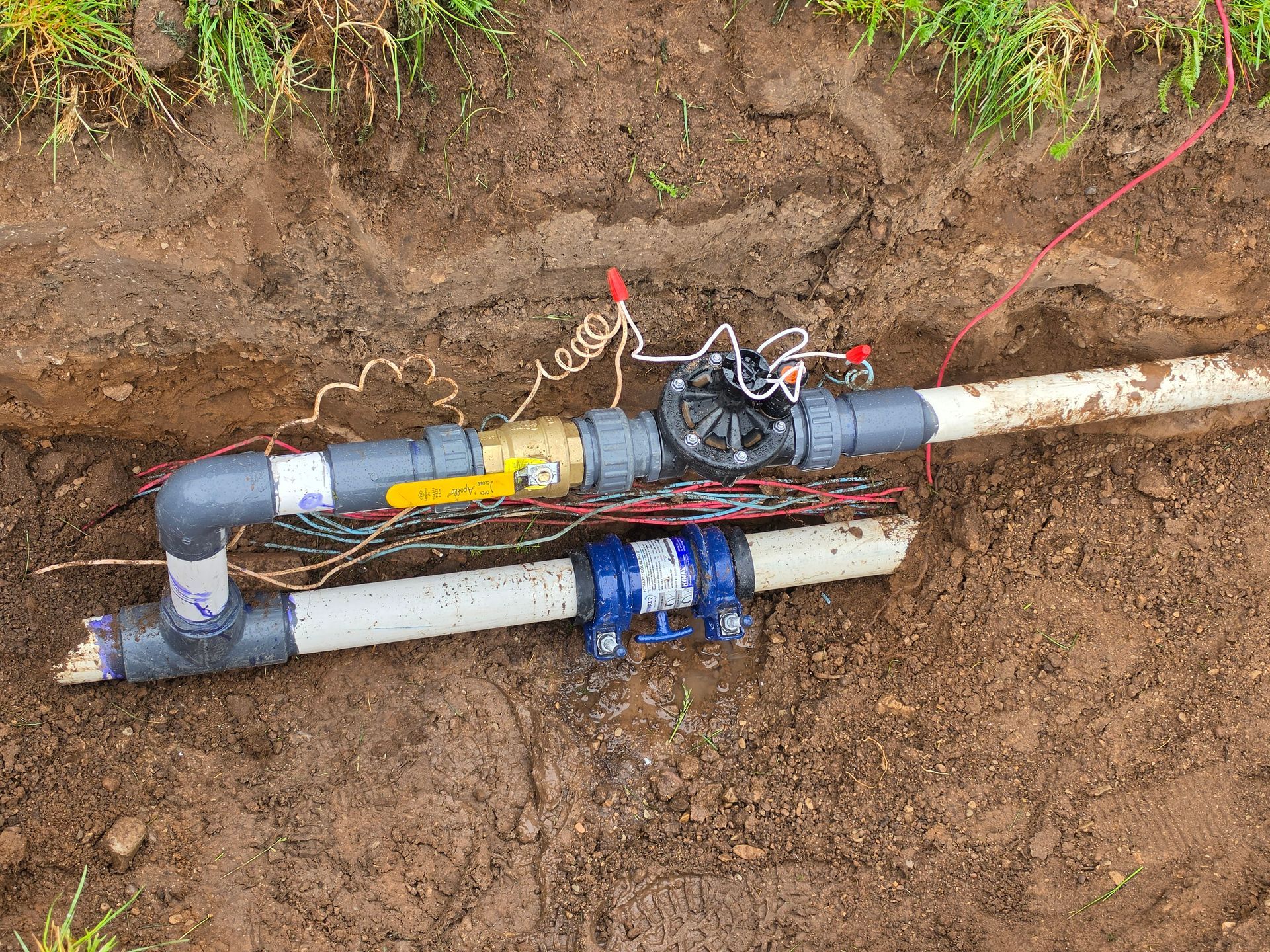 Exposed underground pipe joint with gray and white PVC pipes, red cable, and tools in a dirt trench