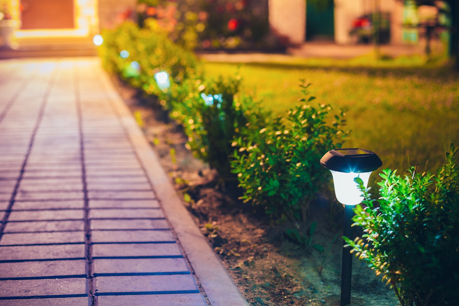 Lit garden path beside trimmed hedges, with glowing walkway lights at dusk.