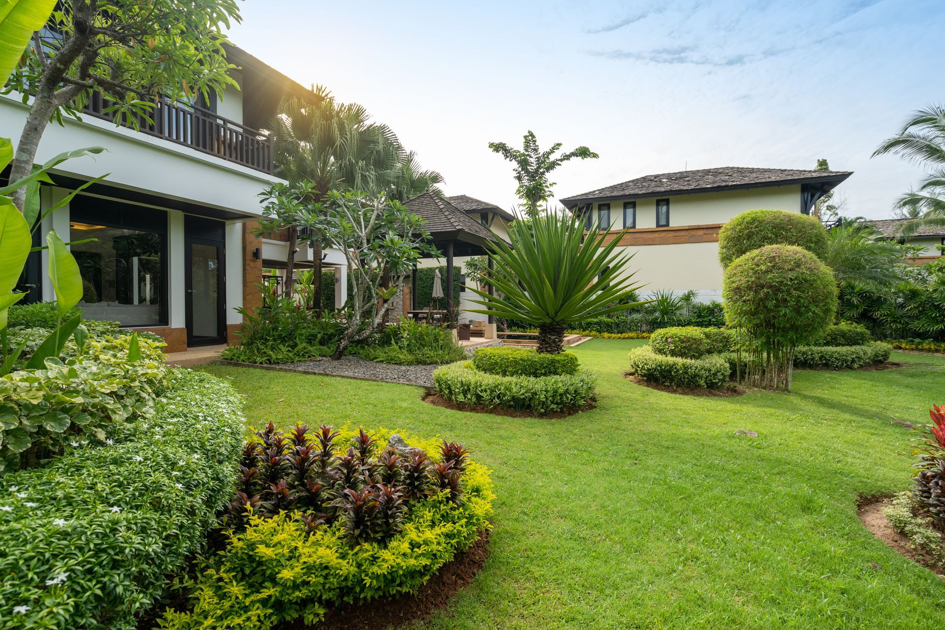 Lush landscaped garden beside modern houses, with green lawns, shrubs, and palm trees under a bright sky
