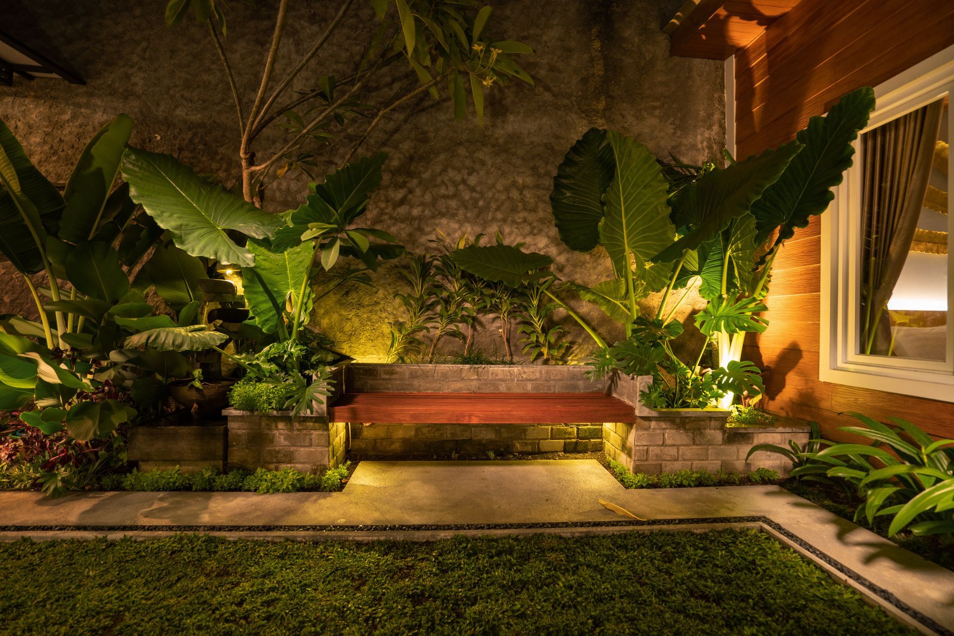 Warm-lit tropical garden with a red bench, lush plants, and stone pathways at night