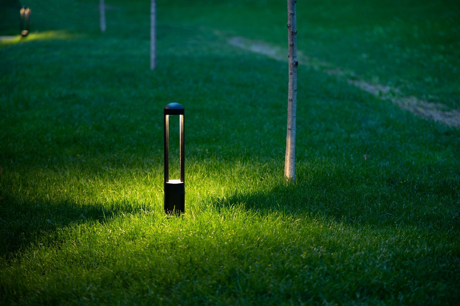 A lit lawn path light glowing on green grass at night, with a nearby pole in the background