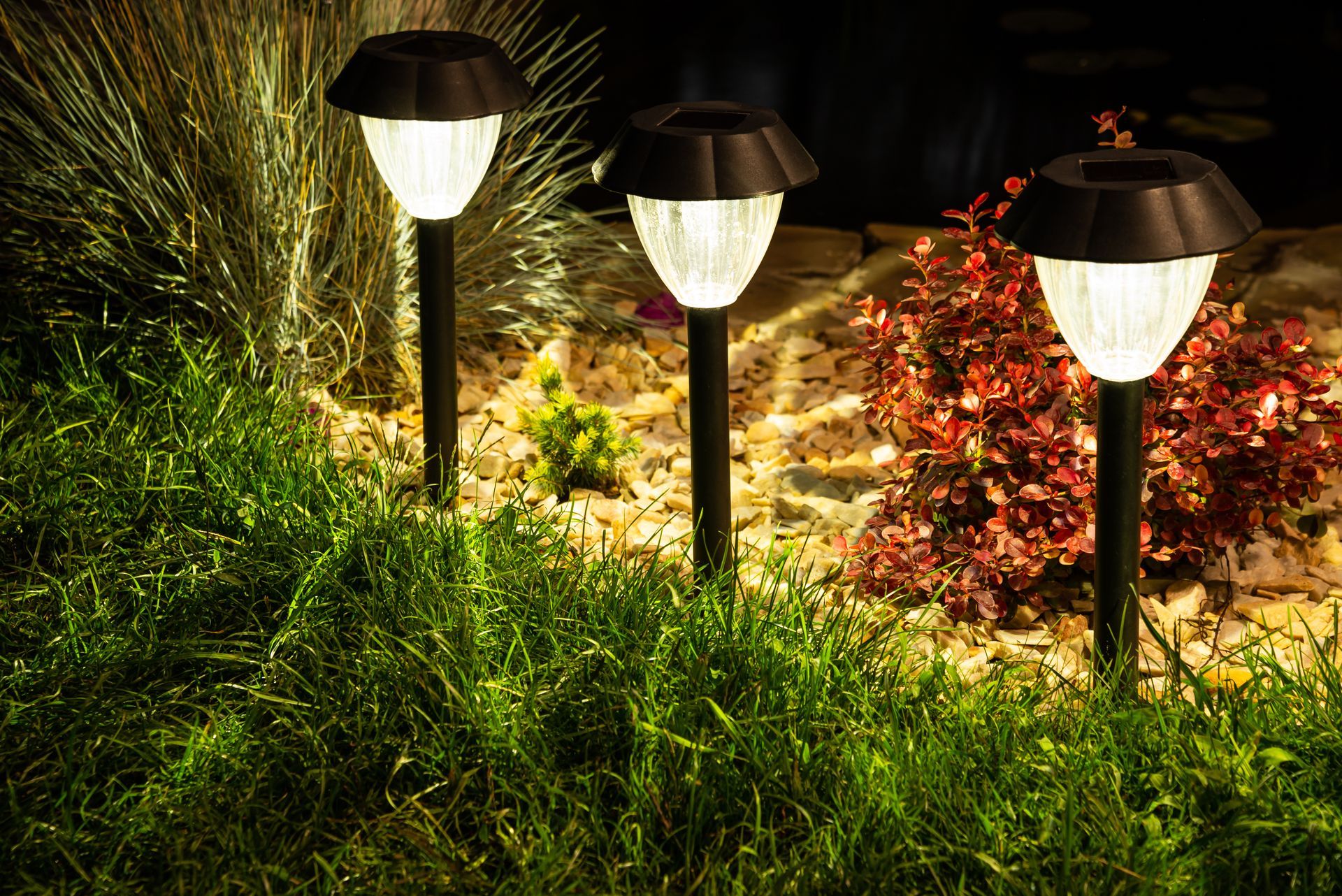 Three lit garden path lamps illuminating grass and flower beds at night