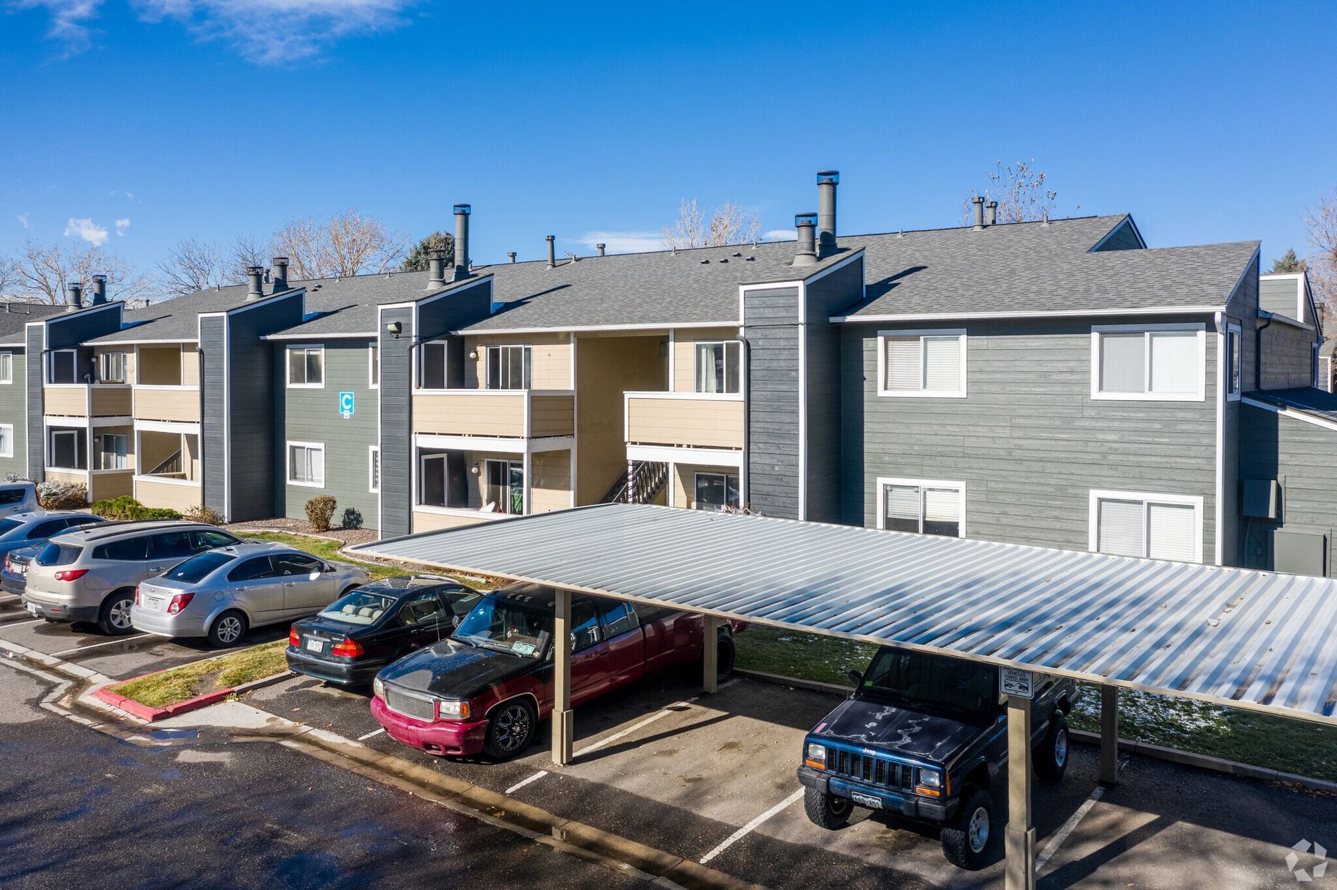 Apartment complex exterior with a carport, parked cars, and blue sky. Buildings are gray and tan at Westward Heights in Denver, CO.