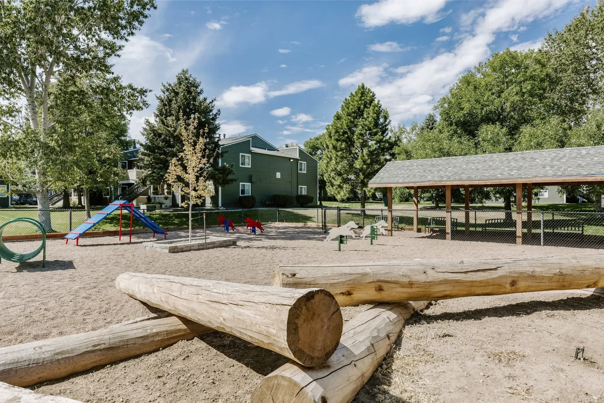 Playground with logs, a slide, and a covered shelter under a bright blue sky with trees and a building in the background at Westward Heights in Denver, CO.