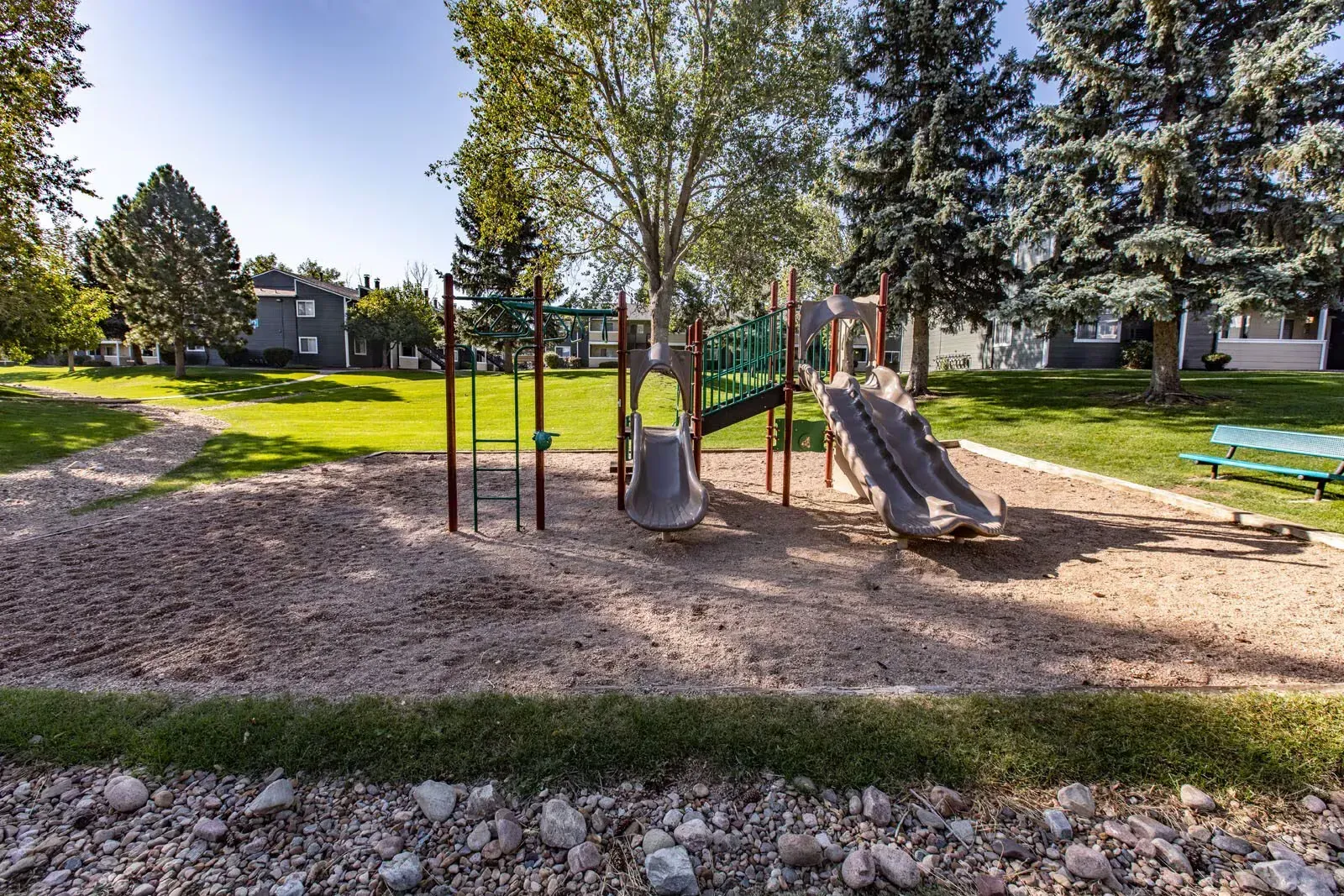 Playground with a slide, climbing bars, and a swing set on wood chips and grass at Westward Heights in Denver, CO.