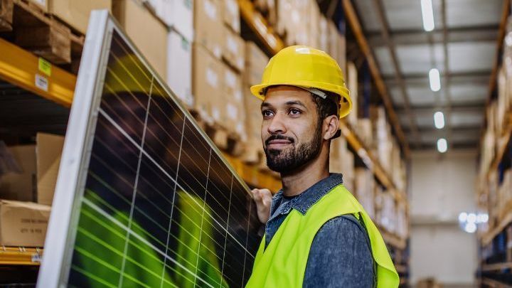 A man wearing a hard hat and safety vest is holding a solar panel in a warehouse.