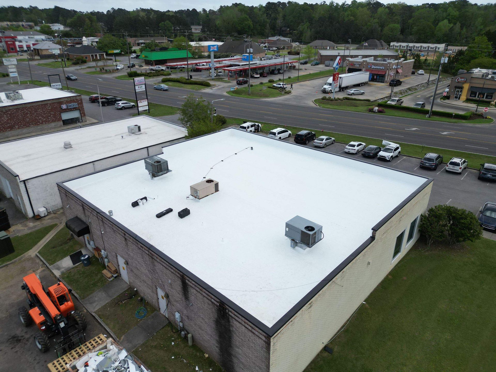 Aerial view of a building with a white roof and parking lot, in a commercial area with cars and green trees.