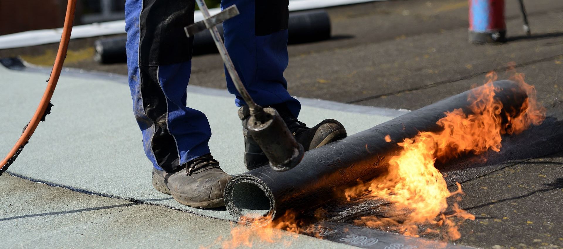 A man is using a torch to burn a roll of roofing material on a roof.