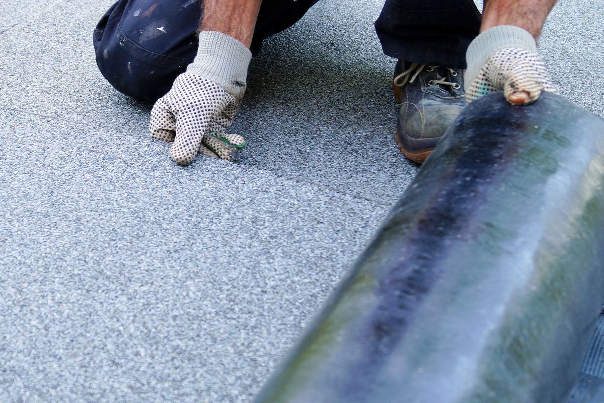 A man is kneeling down on a roof with a roll of roofing material.