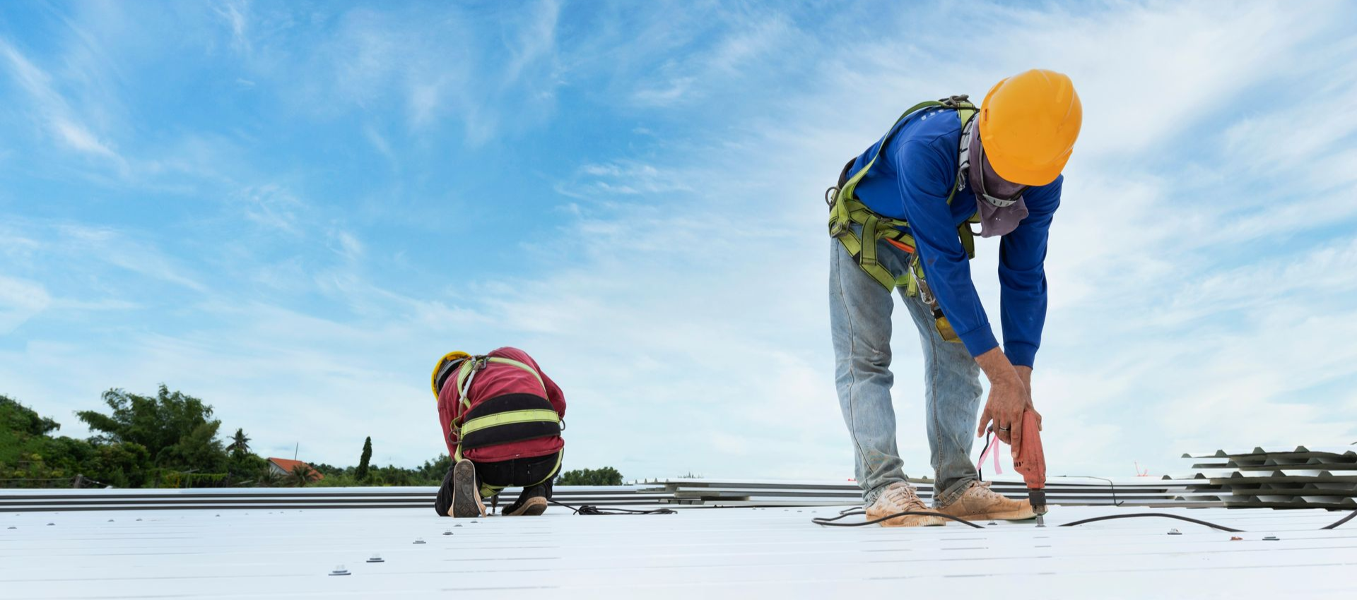 Two roofers in safety gear working on a rooftop under a blue sky.