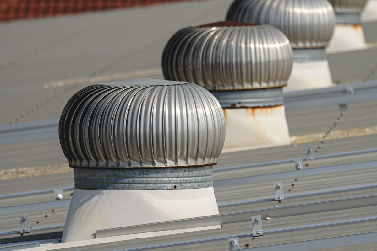 Row of silver turbine vents on a metal roof, with red tile roofs in the background.