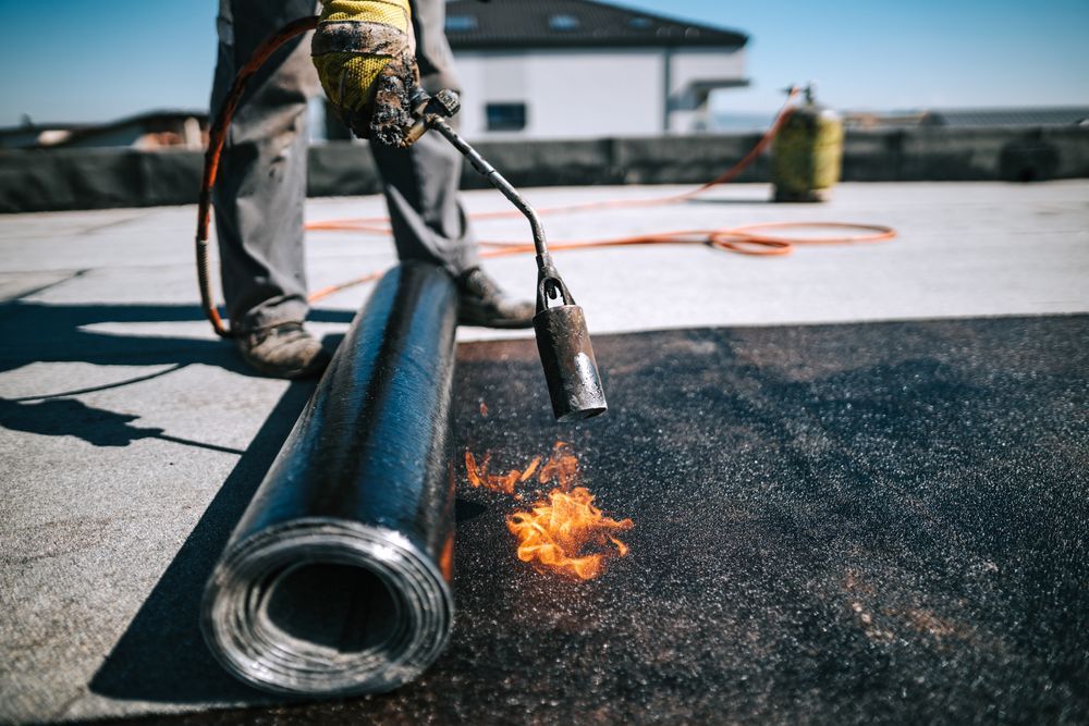 A man is using a torch to burn a roll of asphalt on a roof.