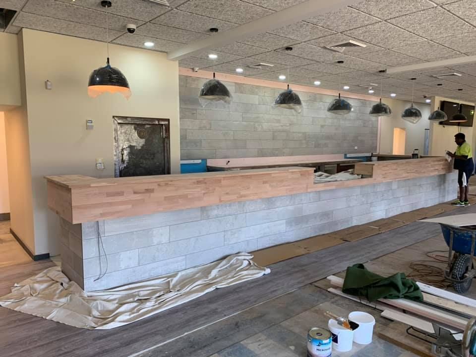 Bar under construction: gray block facade with light wood top, pendant lights, worker at right.