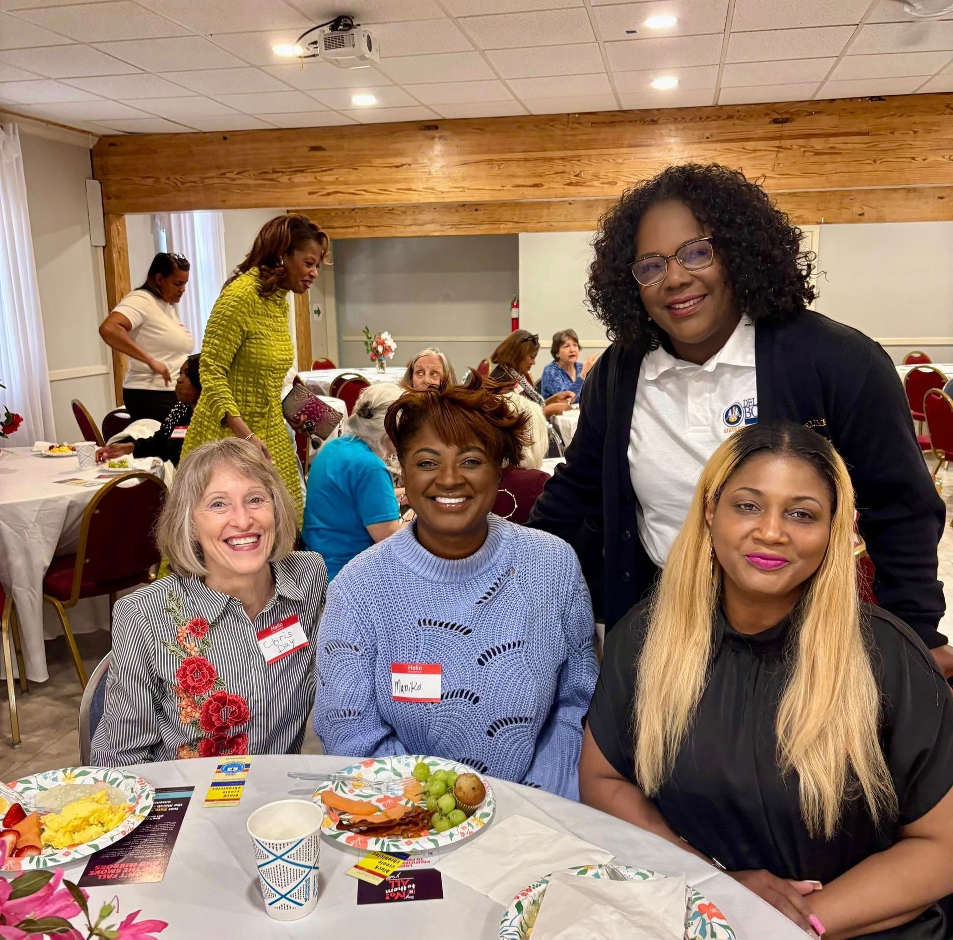 Four women at a table with food, smiling. Others in a room with tables set for a meal.