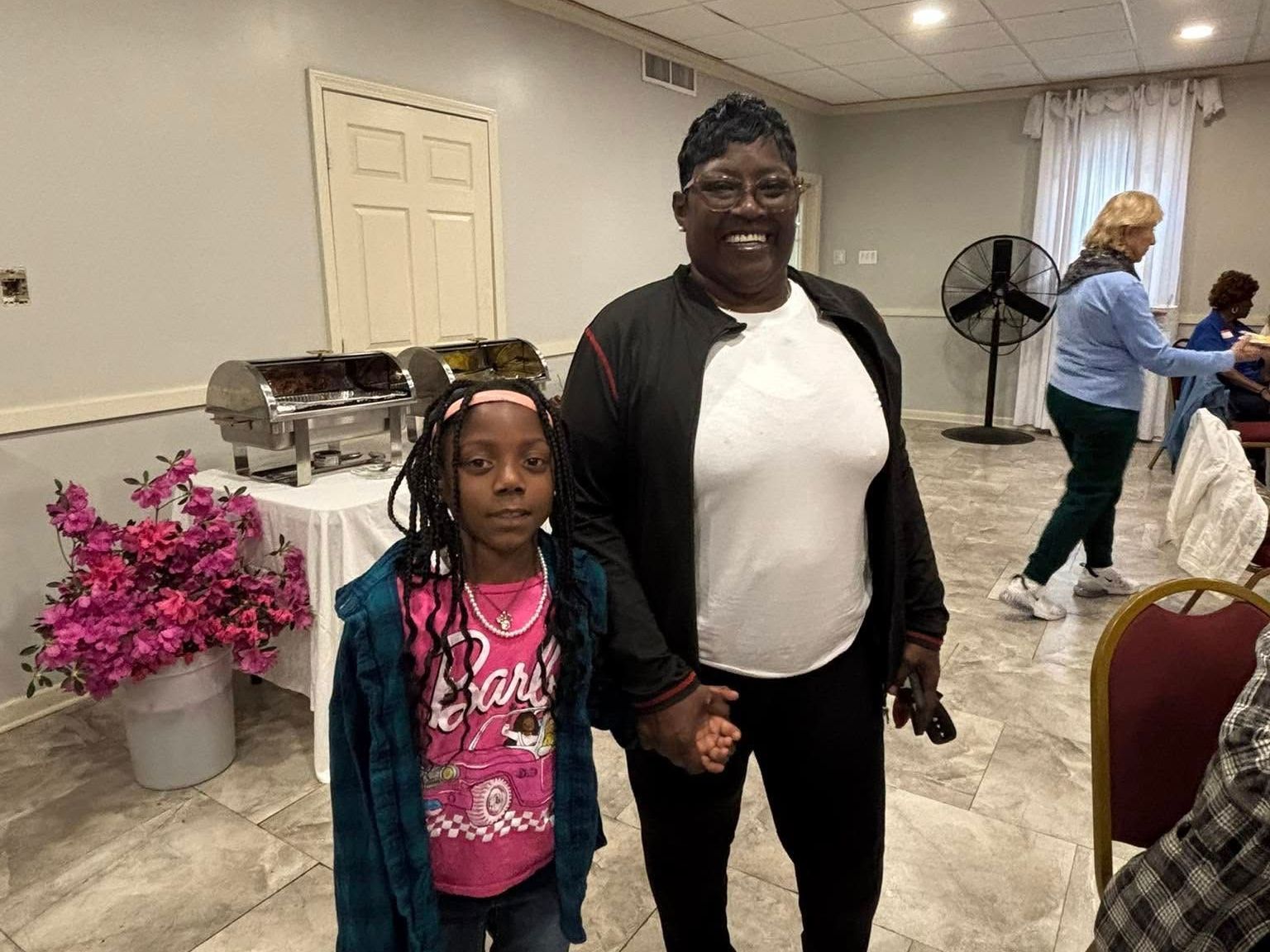 Woman and child standing in front of food table, smiling. Room with decorations and people in the background.