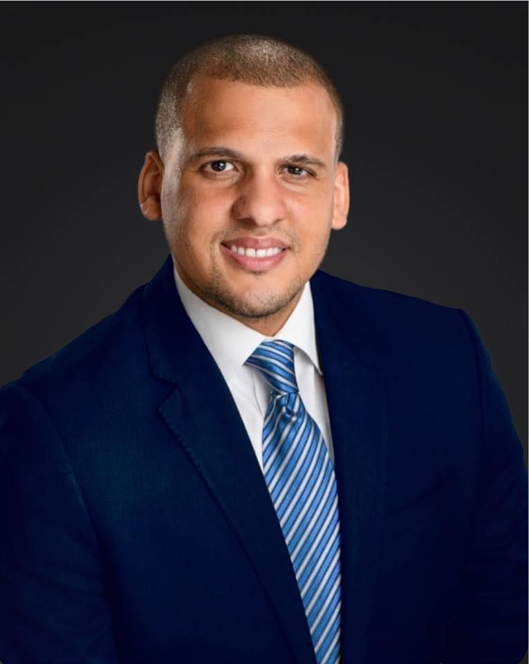 Man in blue suit, smiling, with a striped tie against a dark gray background.