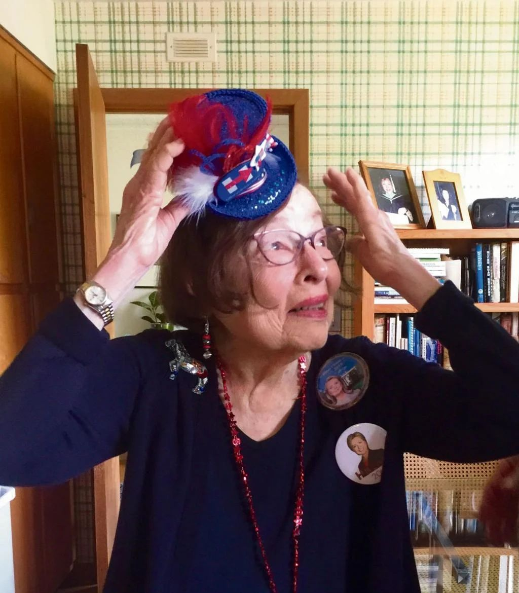 Older person wearing a red, white, and blue hat, hands up, smiling, indoors.