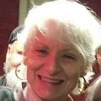 Close-up of a smiling person with short white hair, wearing glasses, and a light-colored shirt.