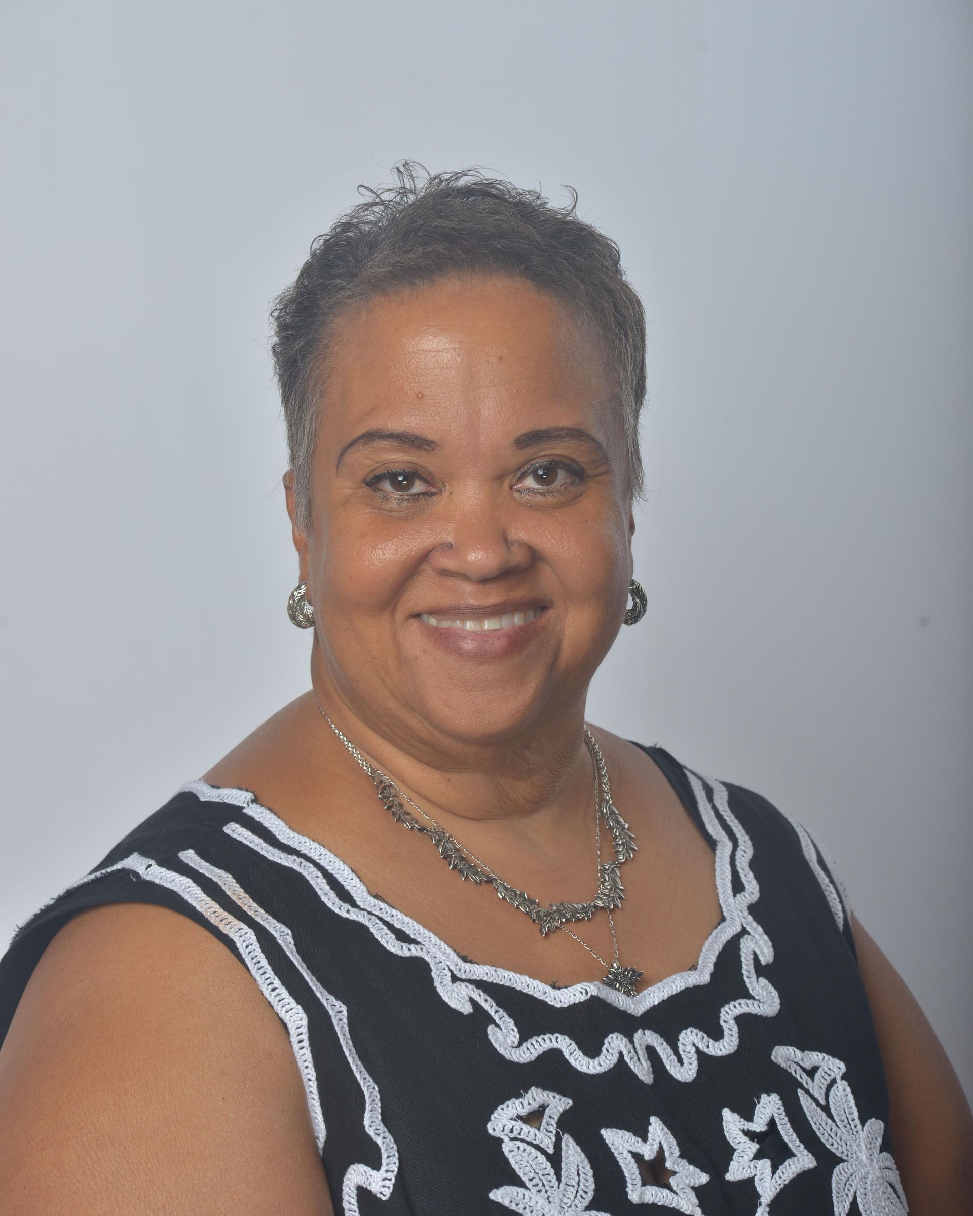 Woman with short hair, smiling. Wearing a black top with white embroidery and a silver necklace.