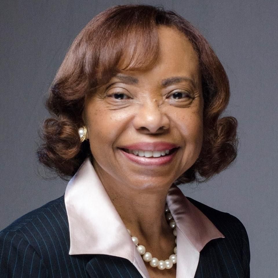 Woman with short brown hair, wearing a pearl necklace, jacket, and smiling.