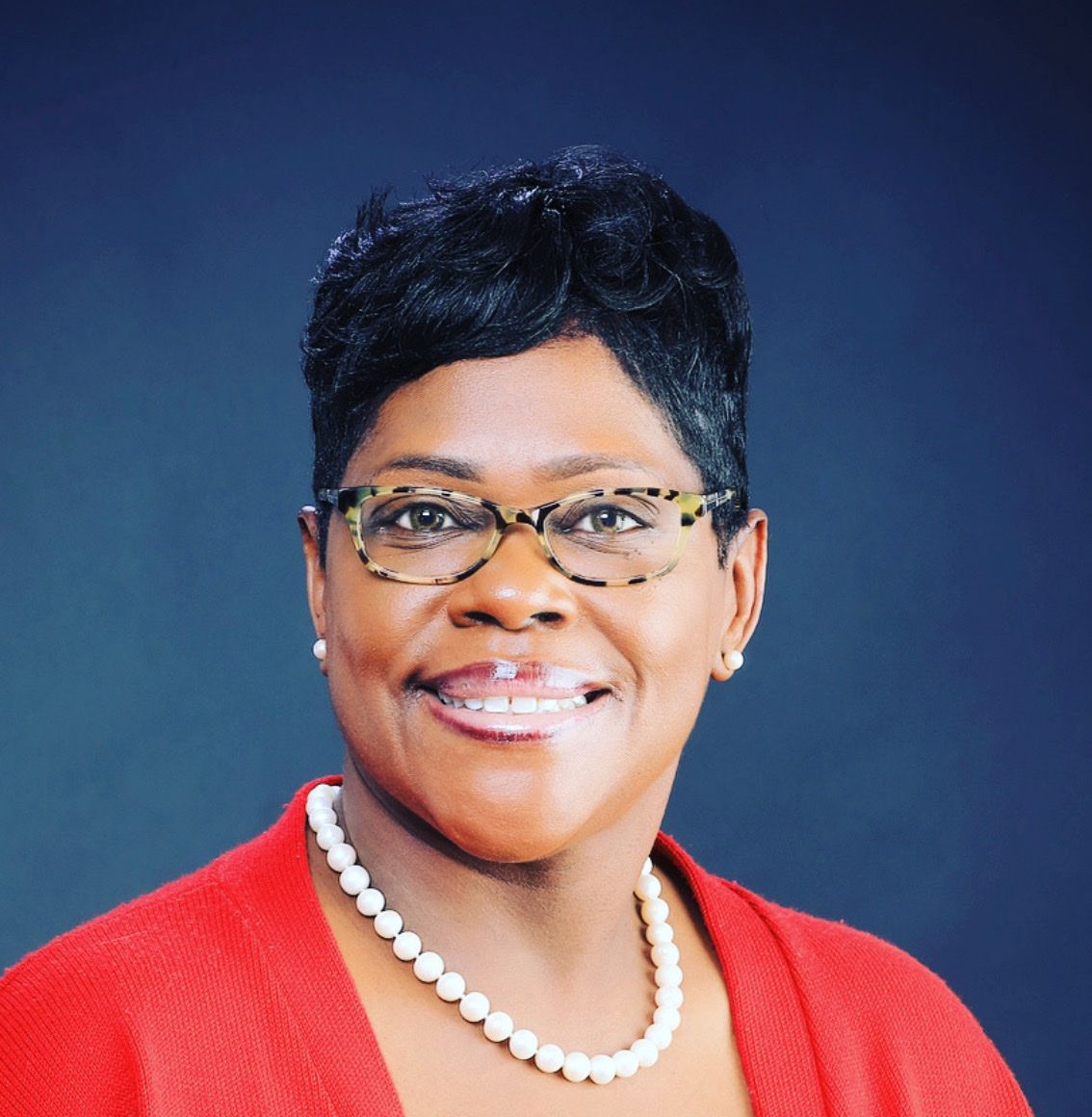 Woman wearing glasses and a pearl necklace smiles, with a red jacket against a blue background.
