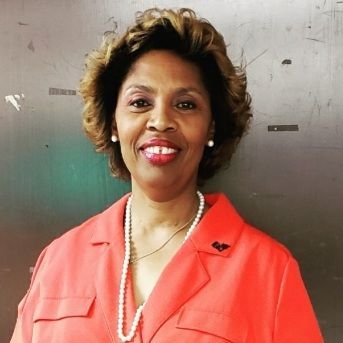 Woman smiling, wearing a coral top, pearl necklace, and small earrings.