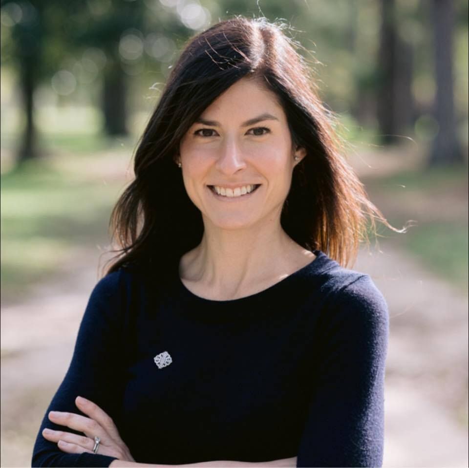 Woman with brown hair smiles, arms crossed, wearing a navy sweater, standing outside with a blurred background of trees.
