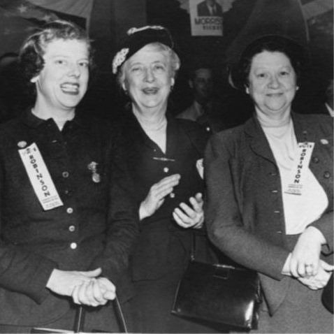 Three women at a political event. One claps, two wear name tags and smile. Dark clothing, dim setting.