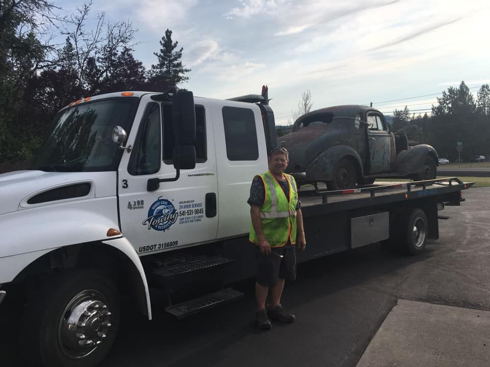A man is standing in front of a tow truck with a car on the back.