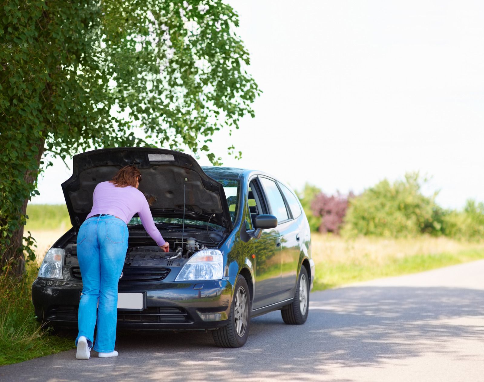 A woman is looking under the hood of a broken down car.