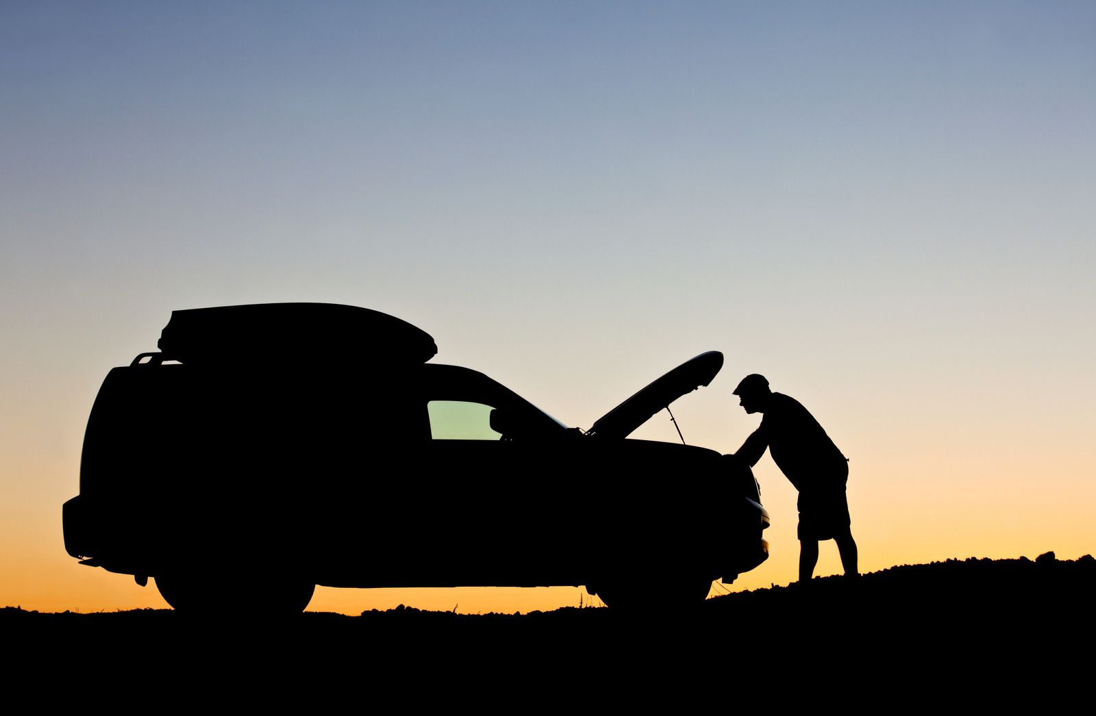 A silhouette of a man looking under the hood of a car