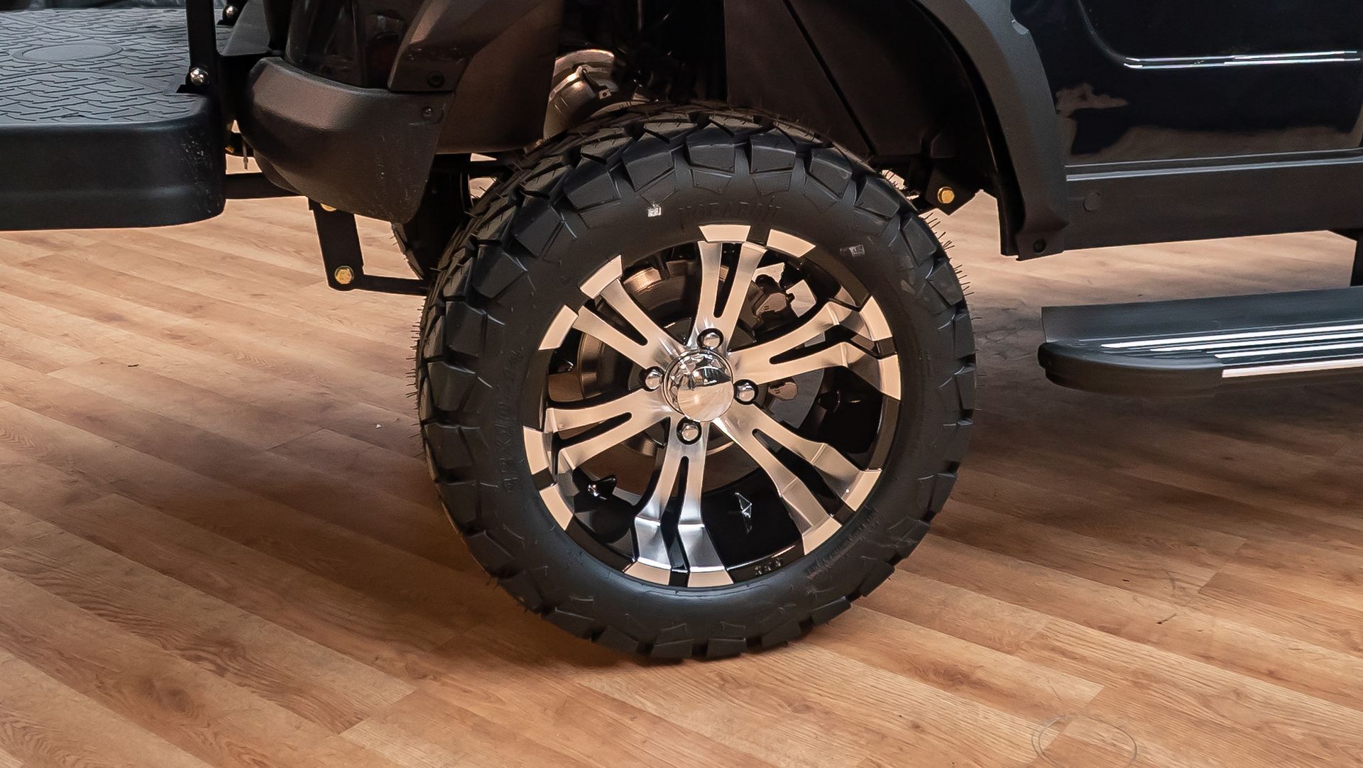 Close-up of a black truck’s large off-road wheel with chrome spokes on a wooden floor.