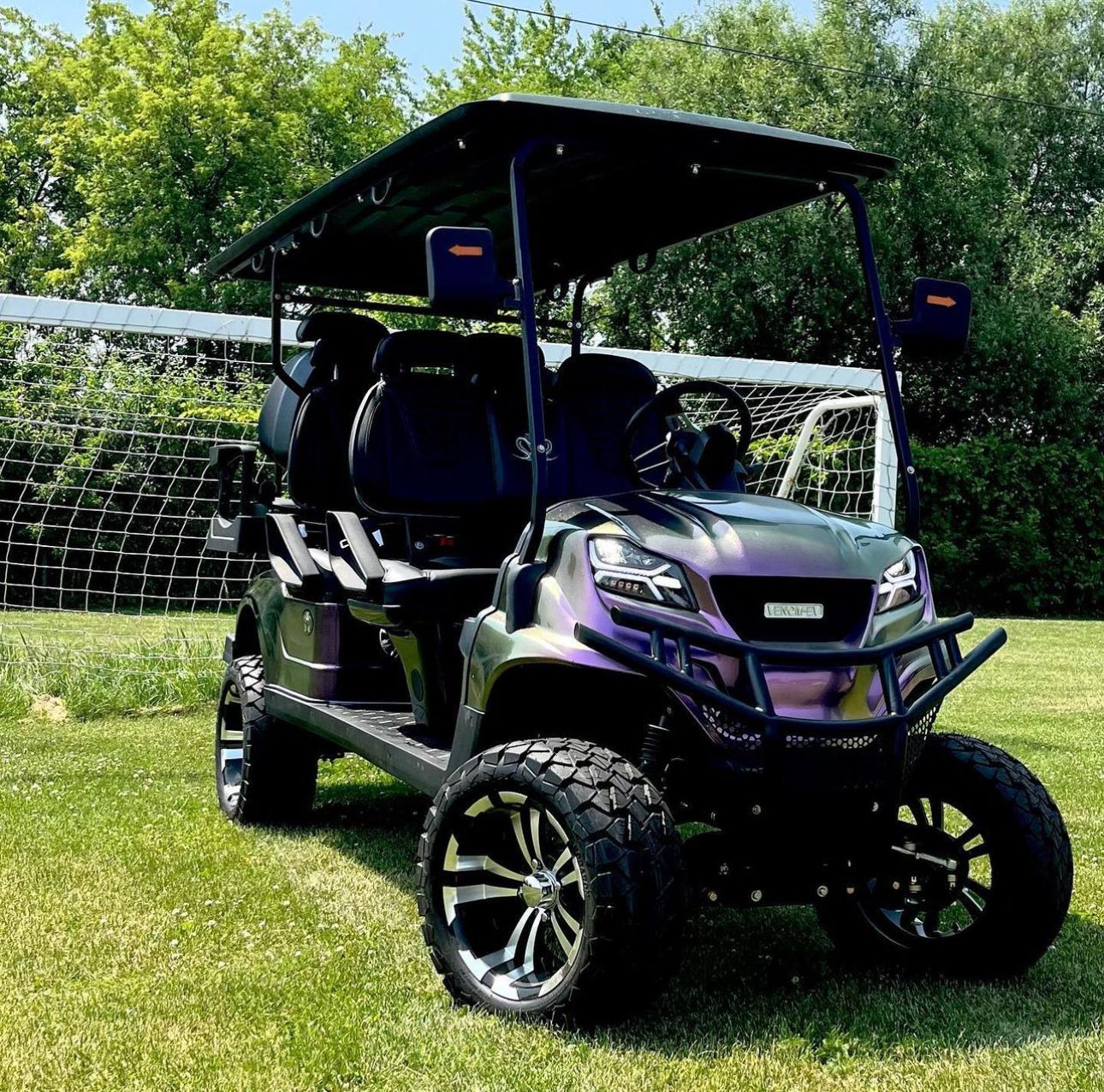 Purple golf cart on grass in front of a netted fence and trees