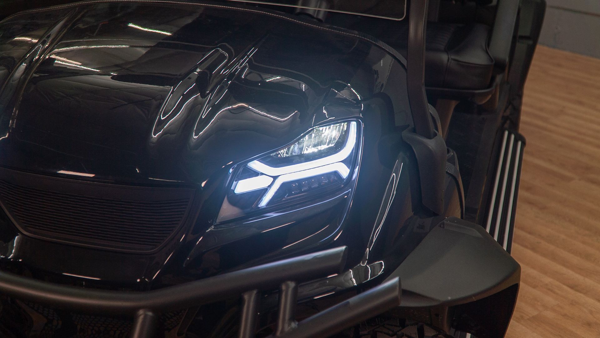 Black toy car with bright headlights on a wooden floor, viewed close-up from the front-left.
