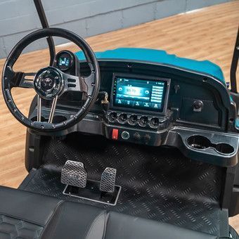 Teal toy car dashboard with steering wheel, screen, pedals, and cup holders on a black floor mat.