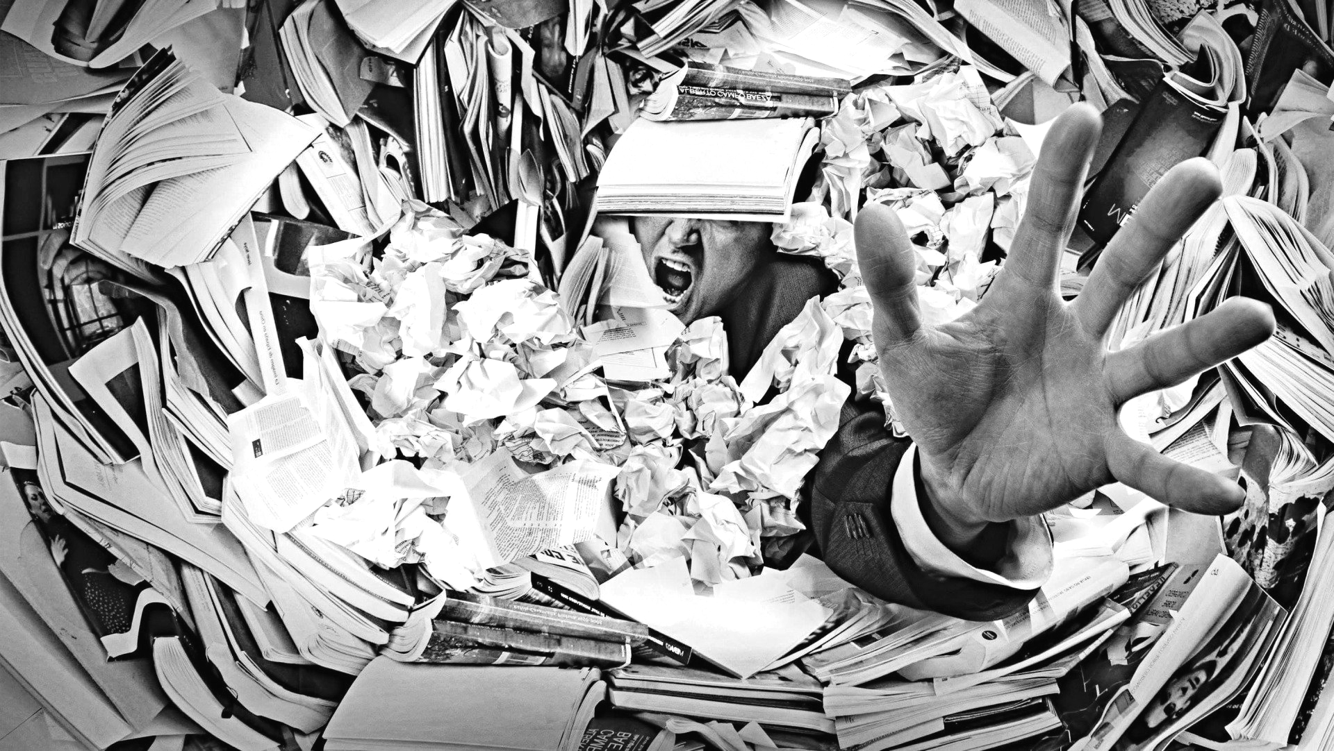 A man in a suit drowning in paper and books, reaching a hand up for help before he goes under