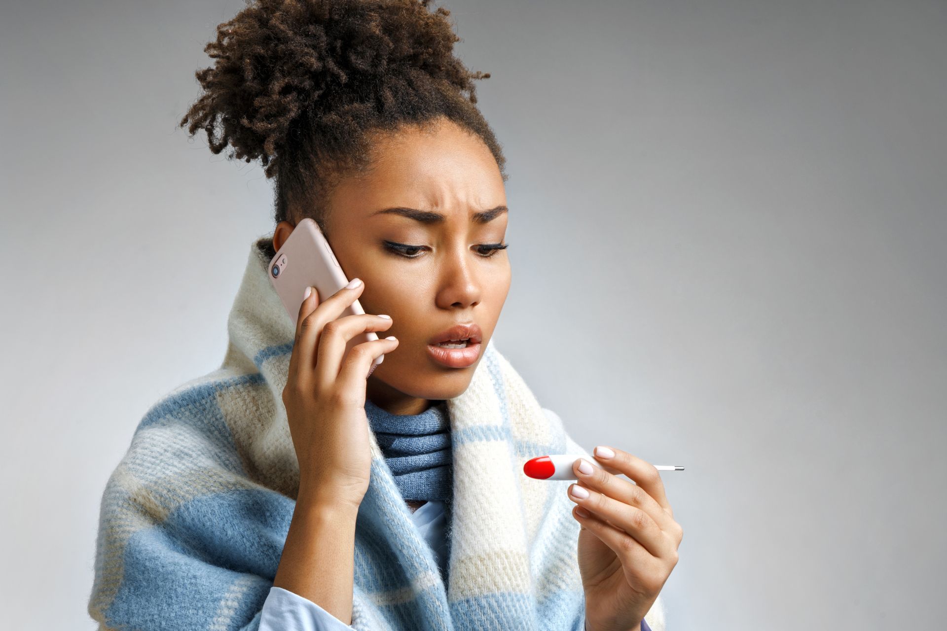 A woman wrapped in a blanket is holding a thermometer and talking on a cell phone.