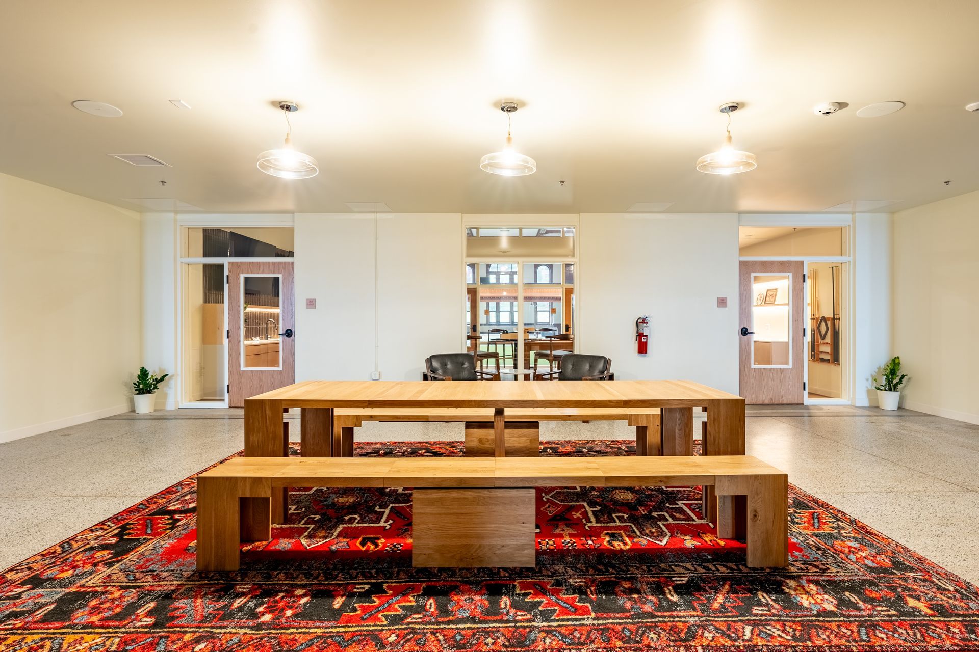 Wooden tables and benches on a large rug in a room with three hanging lights and doors.