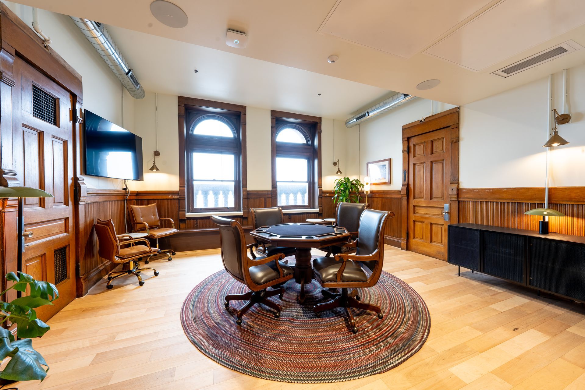 An elegant room with a round poker table, chairs, and wood paneling. Two arched windows and a large rug are present.