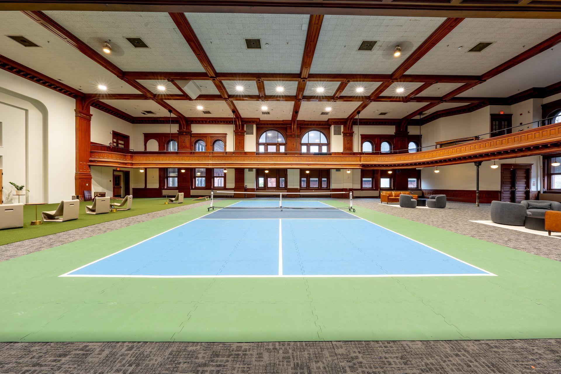 Indoor pickleball court with blue and green flooring, surrounded by seating and a wooden balcony.