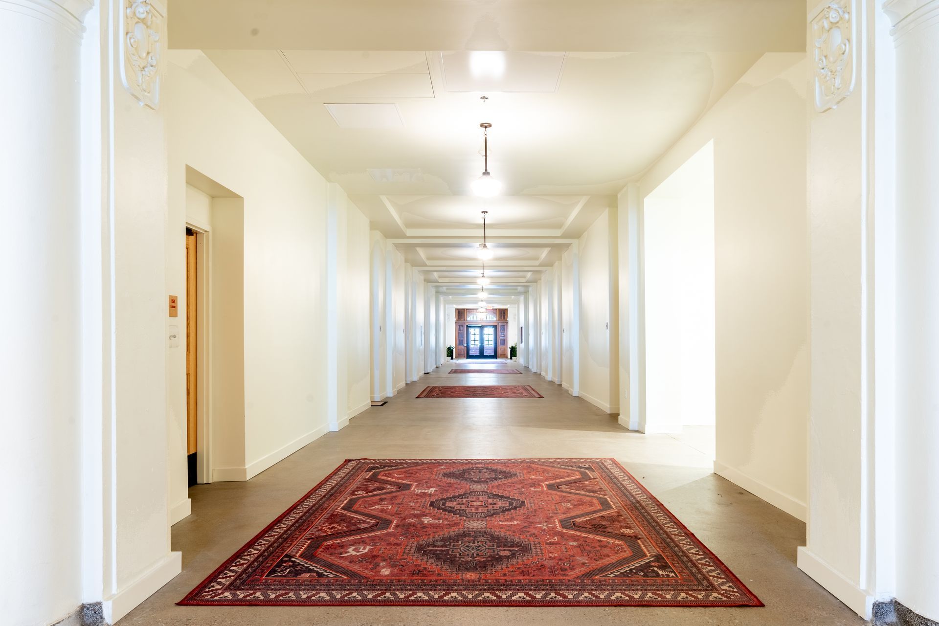 Long hallway with cream walls, Persian rugs, and overhead lights. Doors line the sides.