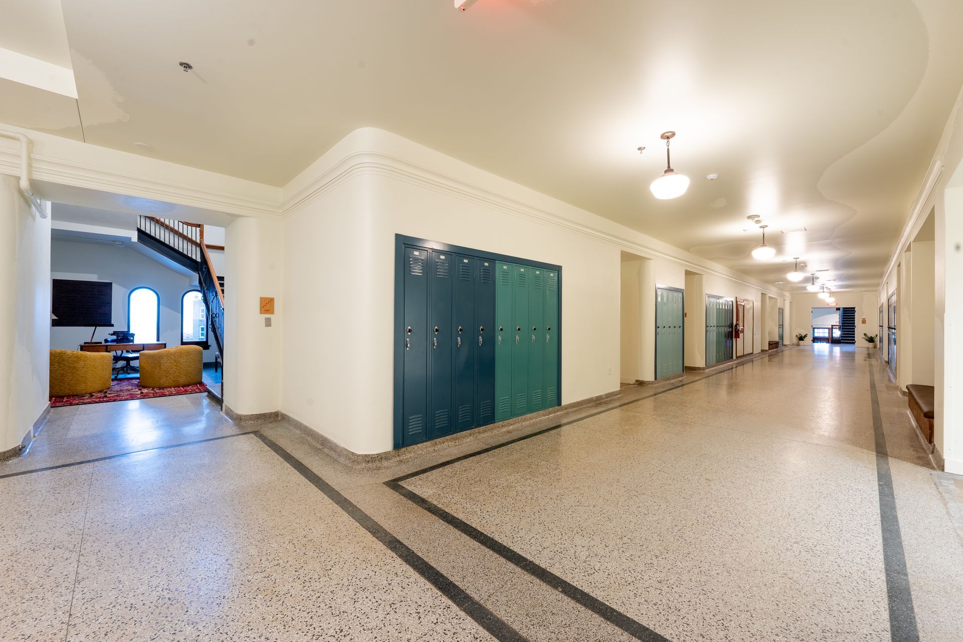 Hallway with lockers, speckled floor, and arched ceiling. A room with furniture is on the left.