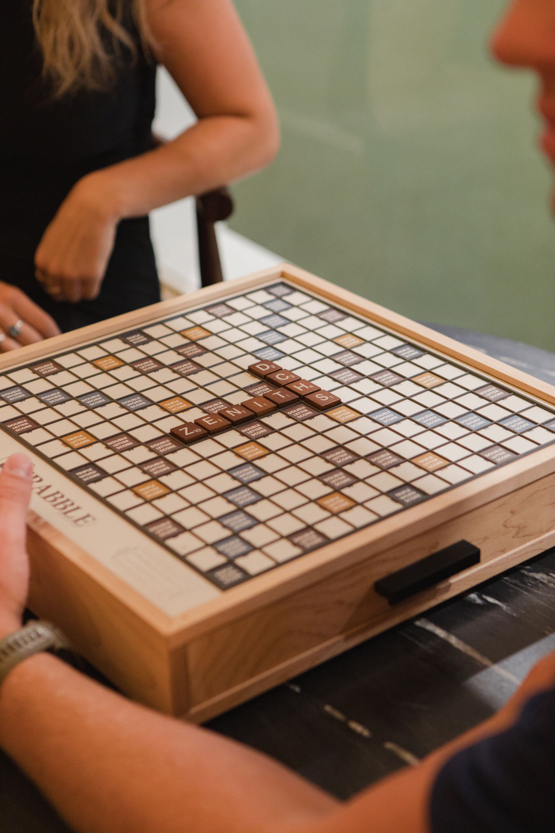 Person playing Scrabble. Wooden game board with tiles, two players visible, seated at a table.