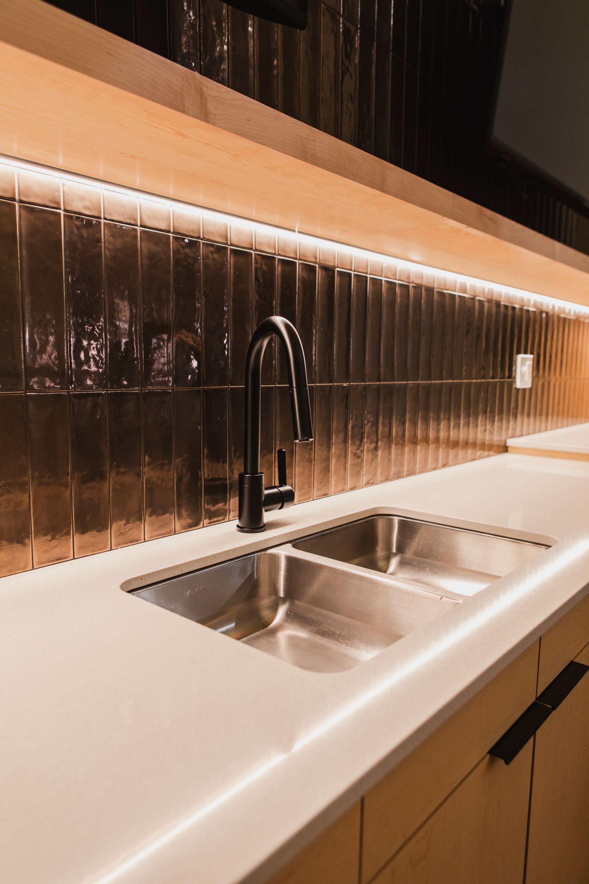 Modern kitchen with stainless steel sink, black faucet, copper-colored tile backsplash, and under-cabinet lighting.