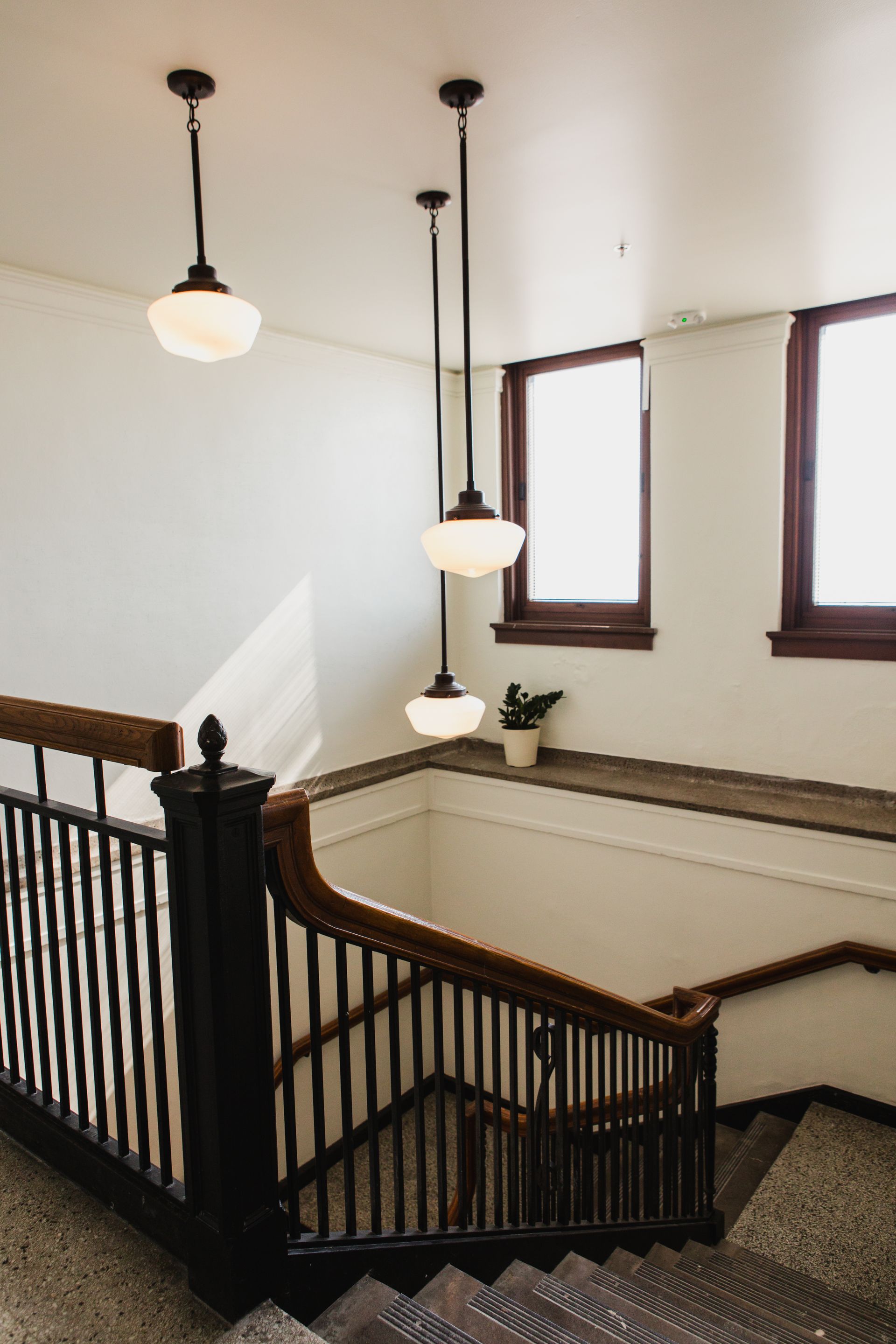 Stairwell with dark wooden railing, white walls, three hanging lights, and windows.