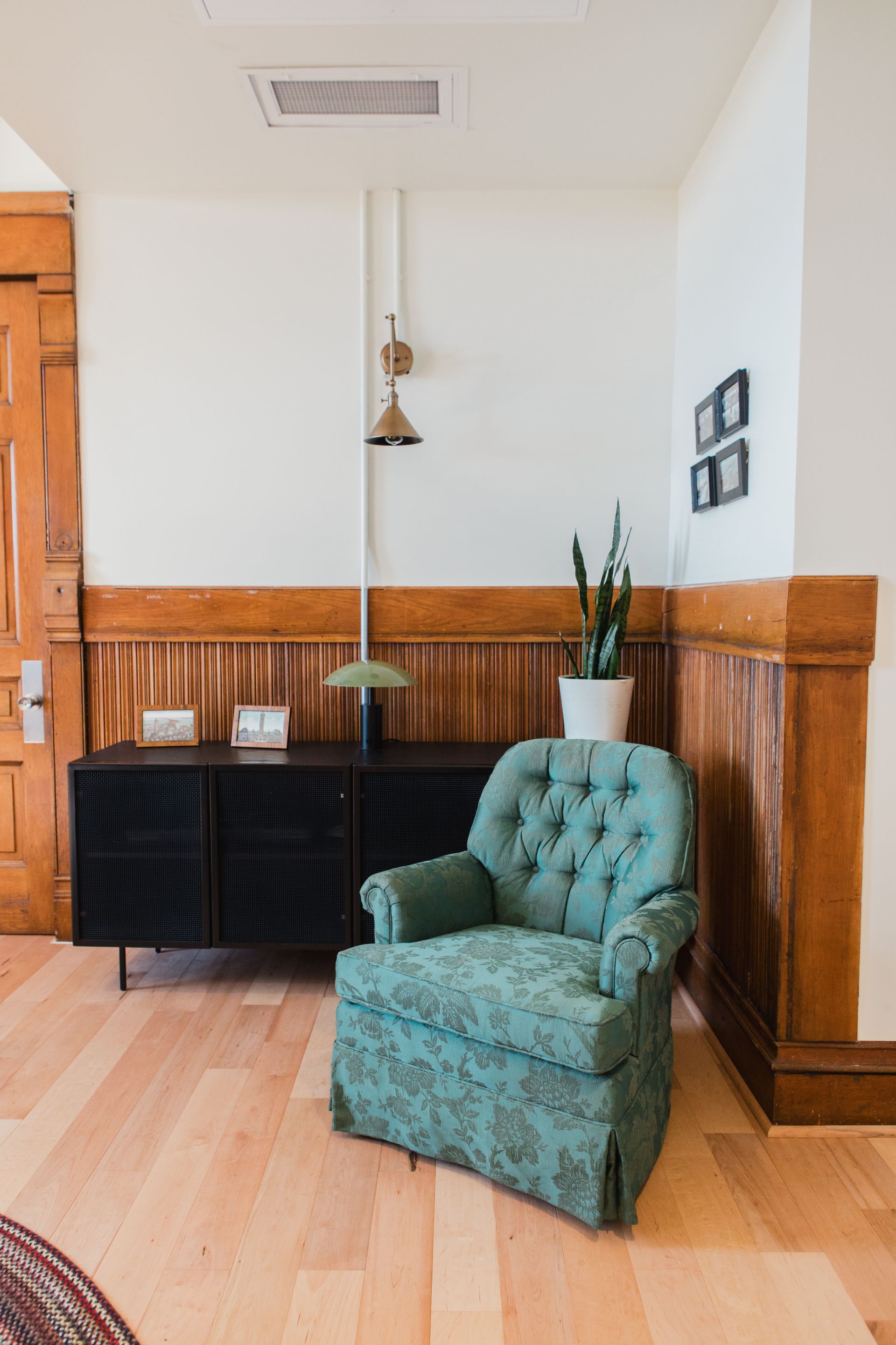 A teal tufted armchair next to a black cabinet, tall cactus, and wood-paneled walls.