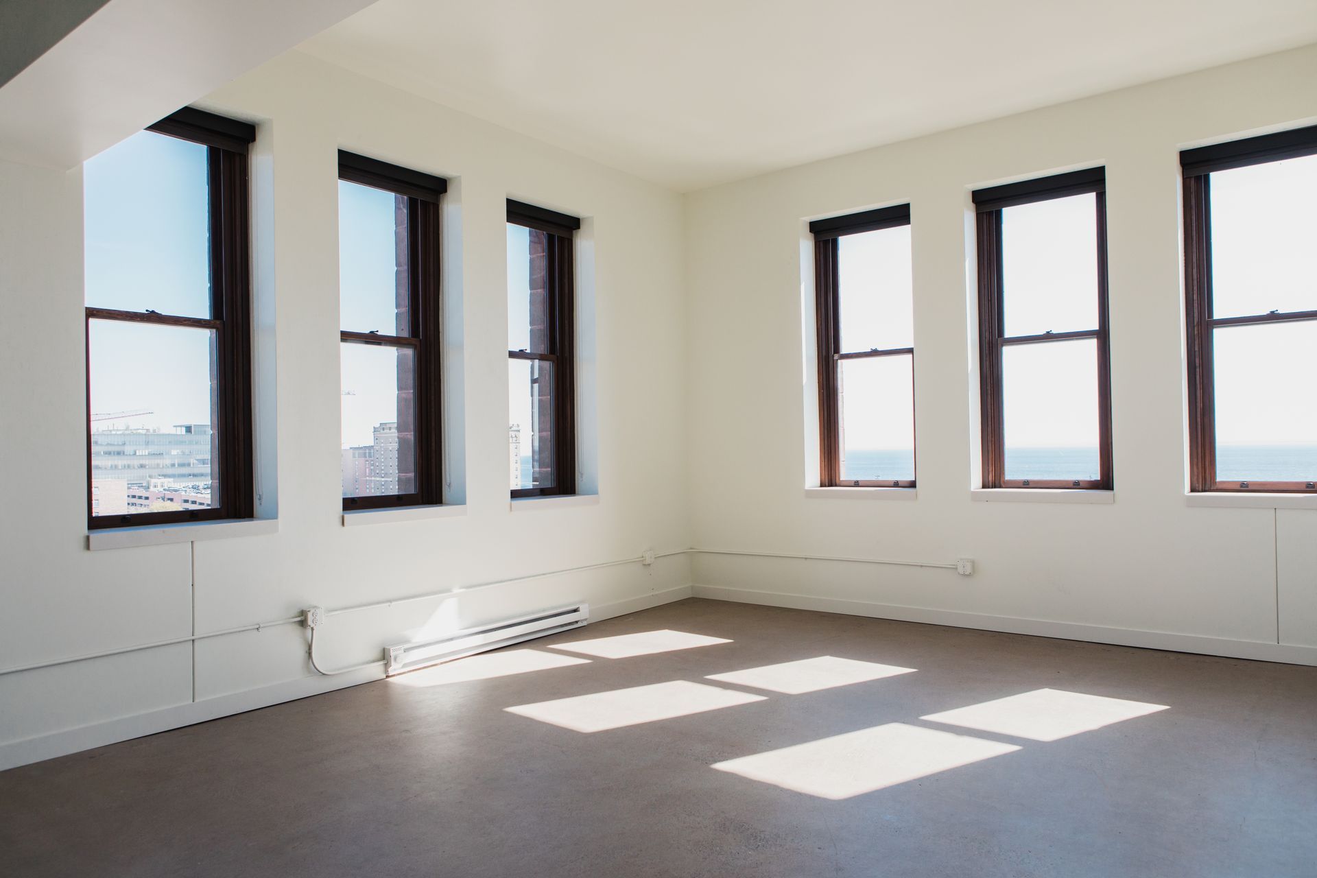 Empty room with large windows, white walls, and dark window frames. Sunlight streams across the floor.