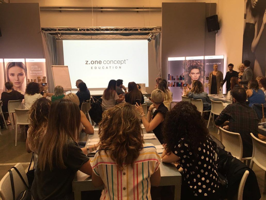 Un grupo de personas están sentadas en mesas frente a una pantalla grande.