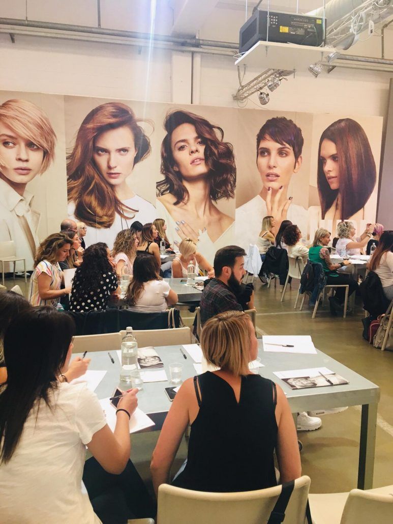Un grupo de personas están sentadas en mesas frente a una pared con fotografías de cabello de mujeres.